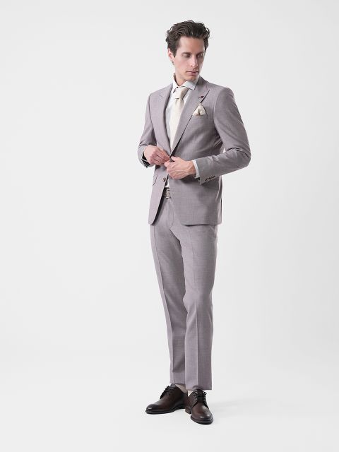 Hombre con traje gris claro, camisa color crema y corbata, de pie ante un fondo blanco sencillo, abotonándose la chaqueta y mirando hacia un lado.