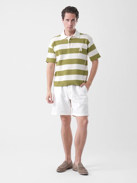 Hombre con polo a rayas verdes y blancas, pantalones cortos blancos y zapatos marrones, de pie frente a un fondo blanco liso.