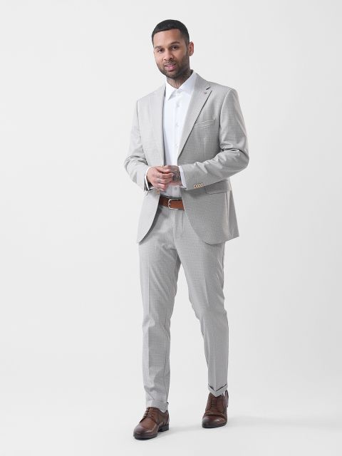 Hombre con un traje gris claro, camisa blanca y zapatos marrones, posando frente a un fondo blanco liso.