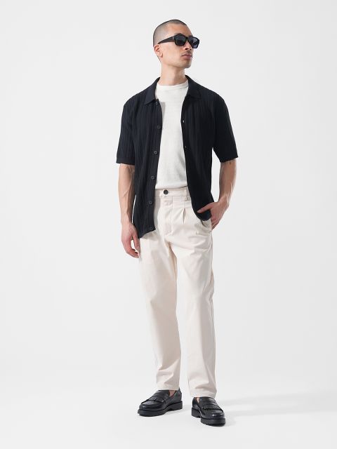 Hombre con gafas de sol, que lleva una camisa negra de manga corta sobre una camiseta blanca, pantalones de color crema y zapatos negros, de pie frente a un fondo blanco.