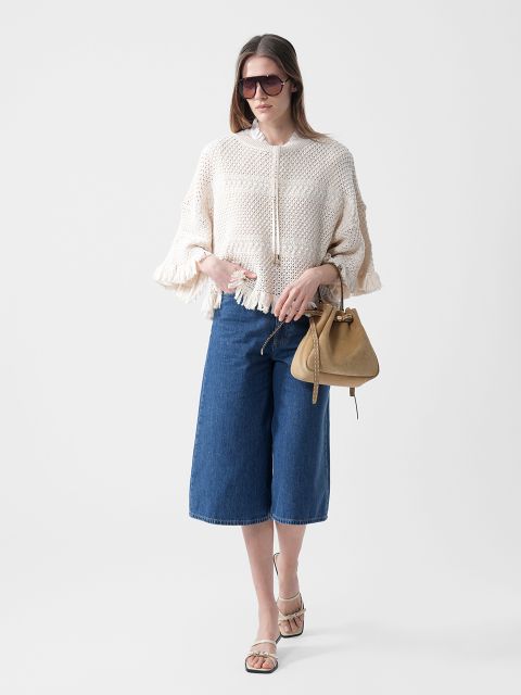 Una mujer lleva gafas de sol, un jersey de punto color crema con flecos, unos pantalones vaqueros culotte azules hasta la rodilla y sandalias, y sostiene un bolso de mano marrón claro sobre un fondo blanco.