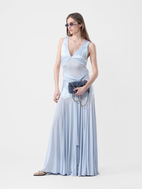 Mujer con un vestido azul claro, fluido y sin mangas, con gafas de sol, sosteniendo un bolso oscuro con cadena en la mano, de pie frente a un fondo blanco liso.