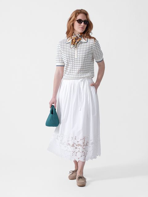 Mujer viste gafas de sol, una parte superior a rayas, una falda midi blanca con bordados, un pañuelo al cuello, un bolso verde azulado y zapatos beige.
