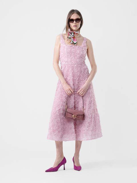 Mujer con un vestido rosa texturizado sin mangas, tacones morados, gafas de sol, un pañuelo alrededor del cuello y con un bolso morado en la mano, de pie frente a un fondo blanco.