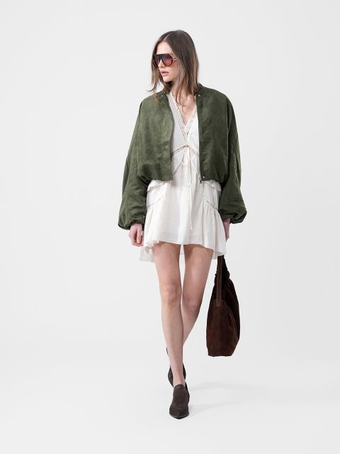 Mujer con vestido blanco, chaqueta verde, gafas de sol y botas marrones, llevando un bolso grande marrón, sobre un fondo blanco liso.