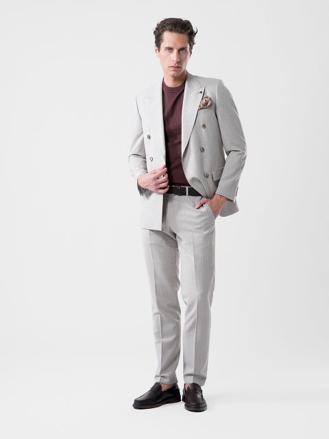 Un hombre con un traje gris claro y una camisa marrón castaño posa con confianza frente a un fondo blanco, con una mano en la chaqueta y la otra en el bolsillo.