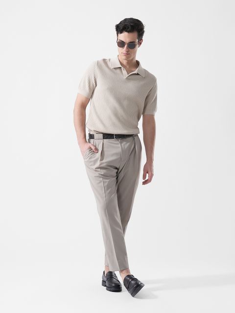 Un hombre que lleva gafas de sol, una camisa de punto de manga corta beige, pantalones de color gris claro y mocasines negros, posa frente a un fondo blanco liso.