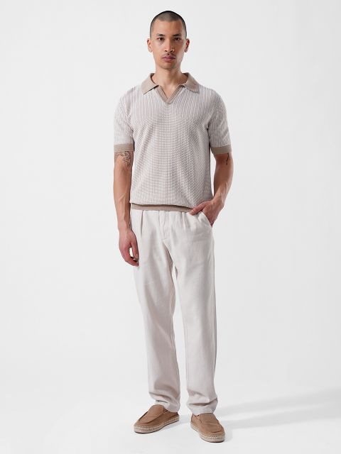 Un hombre está de pie frente a un fondo blanco y lleva una camisa de manga corta de color beige con cuello, pantalones claros y mocasines de color marrón claro.