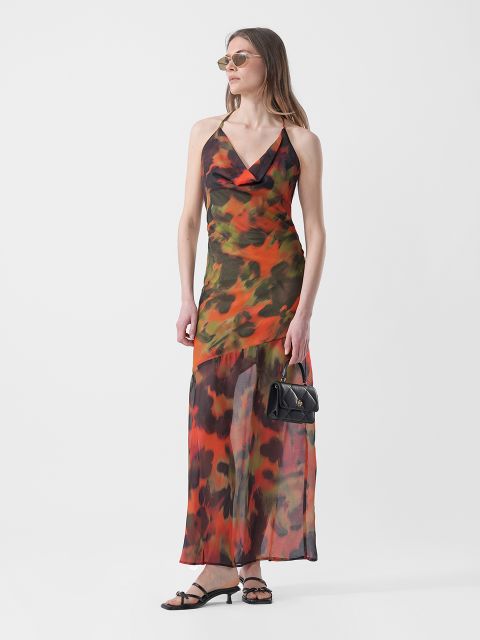 Mujer con un maxi vestido sin mangas con estampado naranja y verde, sandalias y gafas de sol, sosteniendo un pequeño bolso de mano negro, de pie frente a un fondo blanco.