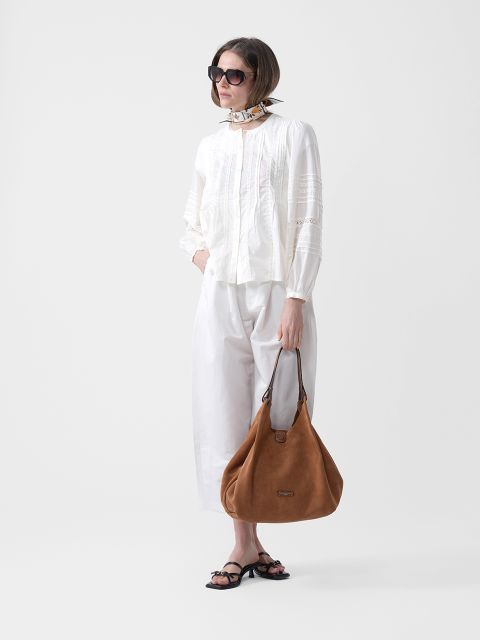 Mujer con gafas de sol, blusa y pantalón blancos, un pañuelo al cuello, sandalias negras y un bolso grande de ante marrón en la mano.