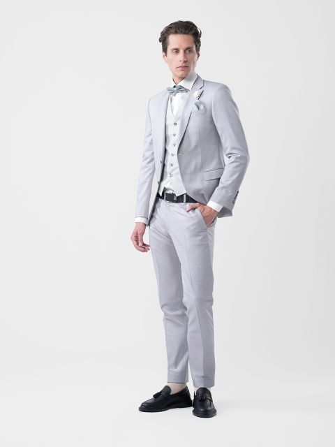 Hombre con traje gris claro, camisa blanca, chaleco estampado y pajarita, de pie frente a un fondo blanco liso.