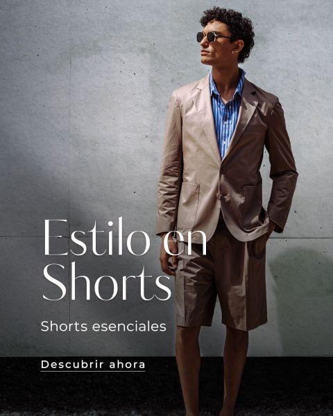 Hombre con gafas de sol, que lleva una chaqueta de traje marrón claro y pantalones cortos a juego sobre una camisa de rayas azules y está de pie frente a una pared gris. El texto dice "Estilo en Shorts.".