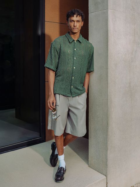 Un hombre con una camisa verde de manga corta, pantalones cortos grises, zapatos negros y calcetines blancos está de pie junto a una pared de hormigón y lleva gafas de sol.