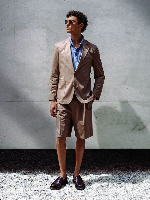 Un hombre que lleva gafas de sol, un blazer marrón claro con pantalones cortos a juego, una camisa de rayas azules y zapatos sin cordones, está de pie junto a una pared de hormigón sobre grava.