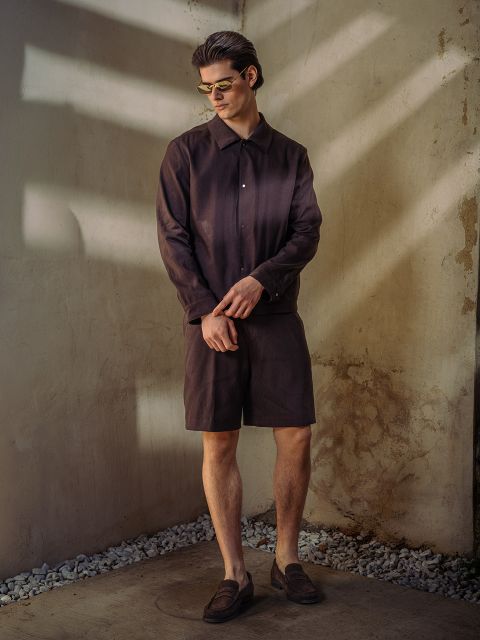Un hombre con gafas de sol, camisa abotonada marrón oscuro, pantalones cortos a juego y zapatos bajos está de pie frente a una pared texturizada iluminada por el sol con detalles de piedra.