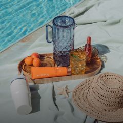 Una bandeja junto a la piscina con cristalería, albaricoques, crema solar, una botella pulverizadora, un sombrero para el sol, una taza blanca y una estrella de mar sobre una toalla verde claro.
