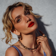 Una mujer con cabello rubio ondulado, un intenso lápiz labial rojo y joyas doradas posa frente a una pared de hormigón, tocándose la nuca y mirando con confianza a la cámara.