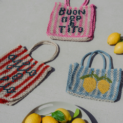 Tres bolsas tejidas de colores, dos con texto en italiano y una con limones, están sobre una superficie clara junto a un plato con limones y albahaca.