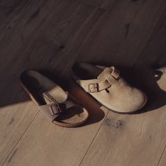 Dos sandalias Birkenstock desparejadas - una de punta abierta y otra de punta cerrada - con hebillas sobre un suelo de madera soleado.