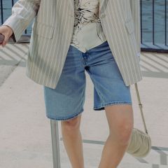 Una persona que lleva un blazer a rayas claras, una blusa estampada y shorts vaqueros, está de pie al aire libre junto a una barandilla y sostiene un pequeño bolso de mano beige.