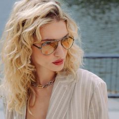 Una mujer con cabello rubio rizado, que lleva gafas de sol de aviador tintadas, un blazer beige a rayas y un collar de cadena grueso, está de pie junto a un cuerpo de agua.