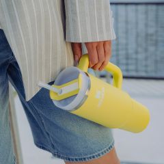 Persona en pantalones cortos de mezclilla y chaqueta a rayas, sosteniendo un gran vaso Stanley amarillo con pajita y asa.