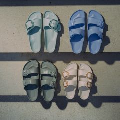 Cuatro pares de sandalias en azul pastel, verde pastel, verde oscuro y blanco roto, dispuestas sobre una superficie de hormigón y atravesadas por sombras.