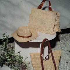Sombrero de paja trenzado y dos sacos de paja con asas marrones sobre una mesa redonda blanca junto a una planta en maceta verde, al aire libre bajo la luz del sol.