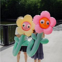Dos personas sostienen grandes flores inflables amarillas y rosas frente a sus caras, mientras están de pie en un camino soleado cerca de una barandilla y de plantas verdes.