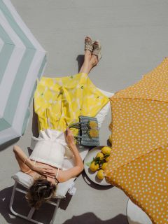 Una mujer con falda amarilla se relaja en una tumbona bajo sombrillas en un día soleado, con limones y un bolso con estampado de limones a su lado.