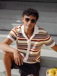 Un hombre con el pelo rizado y gafas de sol está sentado en una escalera exterior, vistiendo una camisa de manga corta a rayas; junto a él hay un cuenco con limones.
