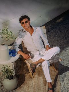 Hombre con gafas de sol y ropa blanca se relaja en una silla al aire libre, junto a una mesa pequeña con vasos azules y un jarrón, sobre una alfombra tejida.