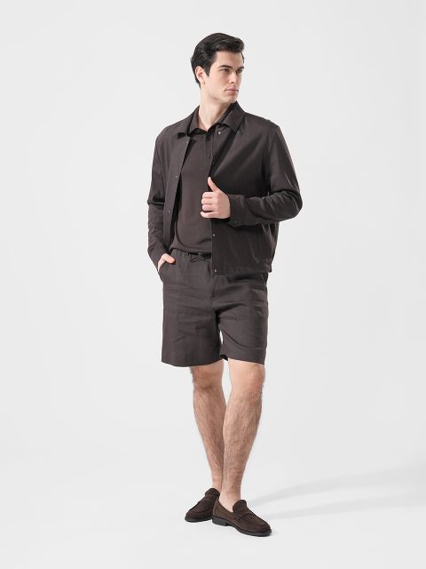 Hombre con chaqueta marrón, polo a juego, pantalones cortos y zapatos sin cordones, de pie frente a un fondo blanco liso.