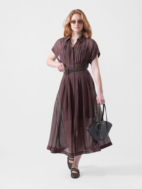 Una mujer con un vestido marrón transparente con cinturón, gafas de sol y sandalias, que lleva un bolso negro, camina con confianza ante un fondo blanco sencillo.