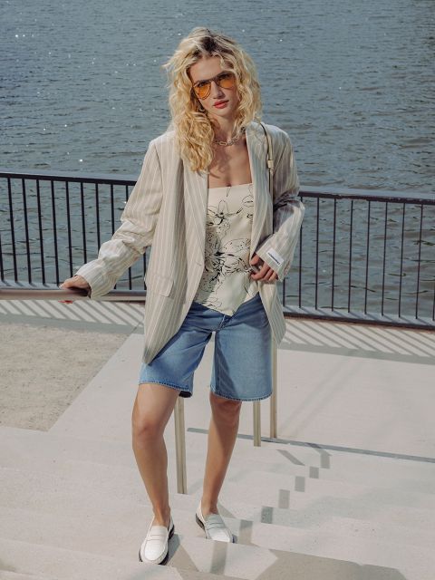 Una mujer de cabello rubio rizado, que viste gafas de sol, un blazer a rayas, una blusa floral, shorts vaqueros y mocasines, está de pie junto a una barandilla a la orilla del agua.