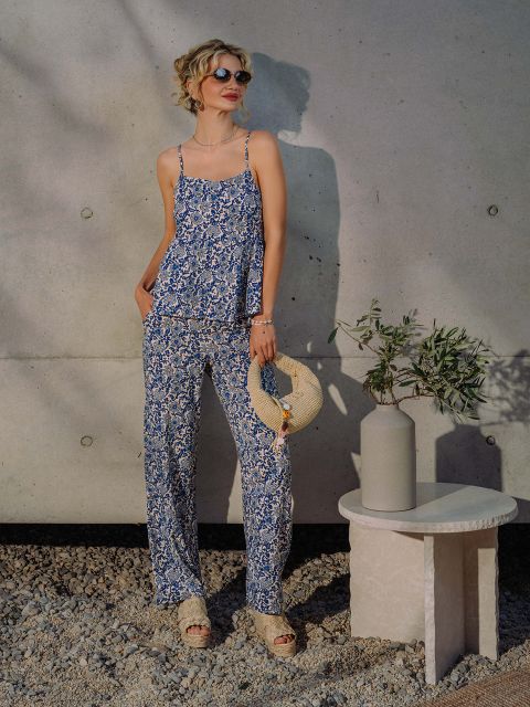 Una mujer con gafas de sol y un atuendo azul floreado está de pie junto a una pared de hormigón y sostiene un sombrero de paja junto a un jarrón con ramas de olivo sobre una mesa pequeña.