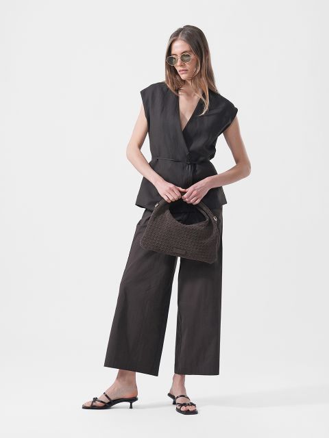 Una mujer con gafas de sol, un top negro sin mangas, pantalones negros de pernera ancha y sandalias de tacón, sostiene un bolso de mano marrón texturizado sobre un fondo blanco.