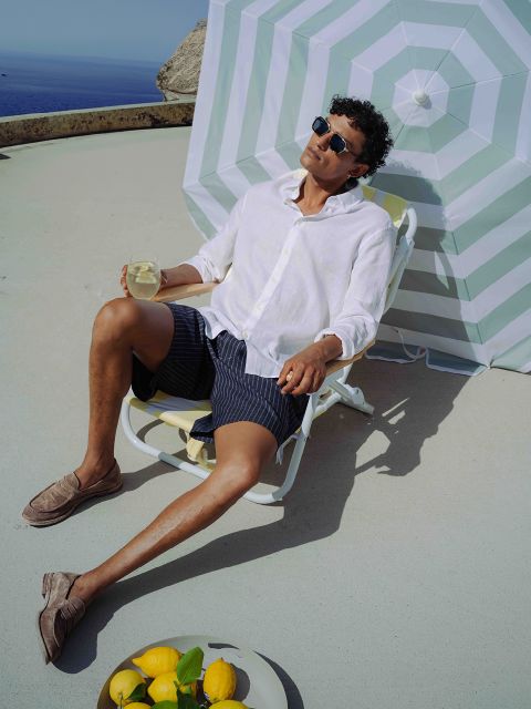 Hombre con gafas de sol y camisa blanca relajándose en una tumbona con una bebida a la sombra de una sombrilla de rayas; limones y un vaso están cerca en el suelo.