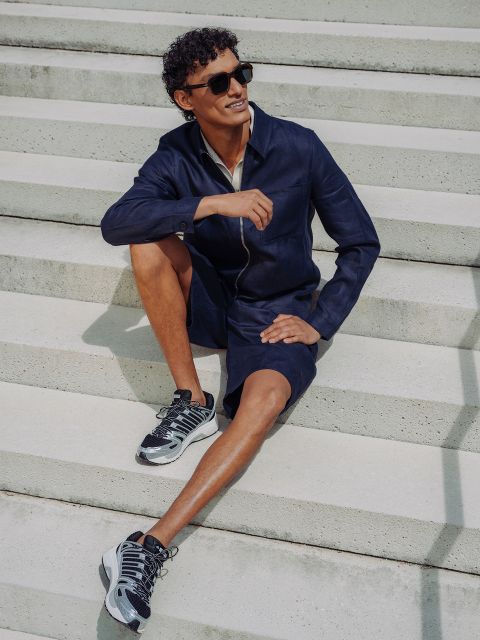 Un hombre con gafas de sol, chaqueta azul marino y pantalones cortos a juego está sentado en una escalera de hormigón al aire libre, sonríe, tiene una pierna doblada y lleva zapatillas deportivas.