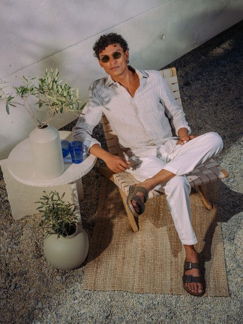 Un hombre con gafas de sol y ropa ligera se relaja en una tumbona junto a una pequeña mesa con plantas y vasos, afuera, sobre un suelo de guijarros y una alfombra tejida.