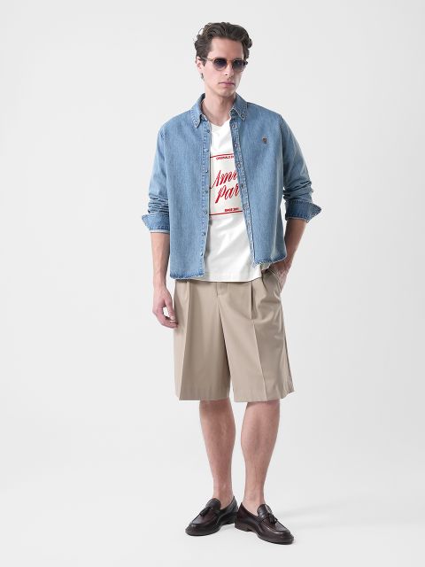 Hombre que lleva gafas de sol, una camisa vaquera sobre una camiseta con gráfico, pantalones cortos beige a la altura de la rodilla y zapatos bajos marrones, de pie frente a un fondo sencillo.