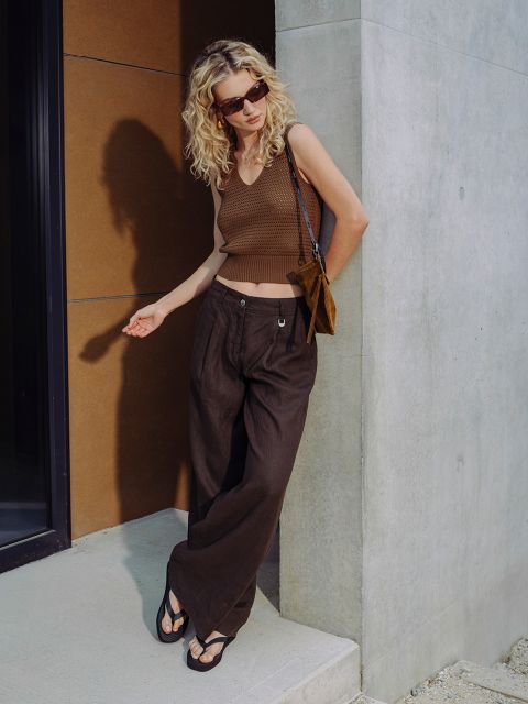 Una mujer con cabello rubio rizado y gafas de sol lleva un top sin mangas marrón, pantalones anchos y sandalias, y está de pie frente a un edificio moderno.