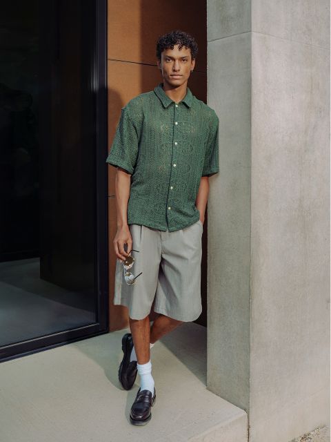 Un hombre con una camisa verde de manga corta, pantalones cortos grises, calcetines blancos y zapatos negros, apoyado en una pared y sosteniendo unas gafas de sol en una mano.