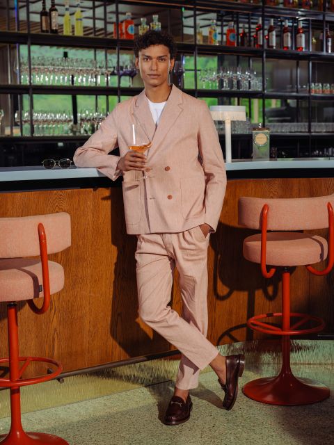 Un hombre con un traje rosa claro sostiene una bebida en la mano mientras se apoya en una barra de bar, detrás de la cual hay taburetes de bar rosas y estantes con vasos y botellas.