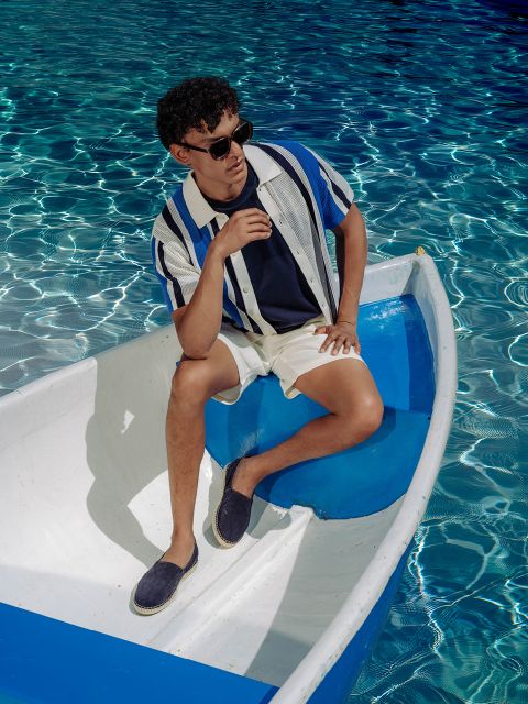 Un hombre con gafas de sol, camisa a rayas y pantalones cortos está sentado relajadamente en un pequeño bote blanco y azul sobre el agua azul clara y brillante.