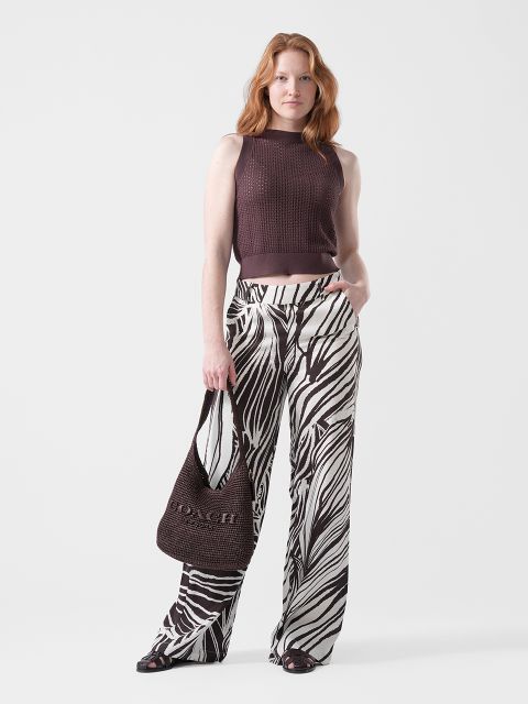 Una mujer lleva una blusa marrón sin mangas, unos pantalones anchos con estampado de cebra, sandalias negras y un bolso Coach marrón.
