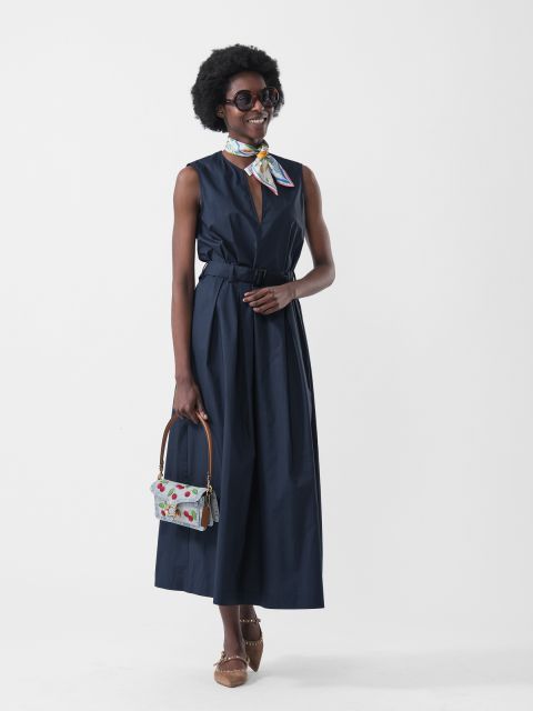 Mujer con un vestido azul marino sin mangas, una bufanda colorida, gafas de sol, un bolso estampado y zapatos marrones, sobre un fondo blanco.
