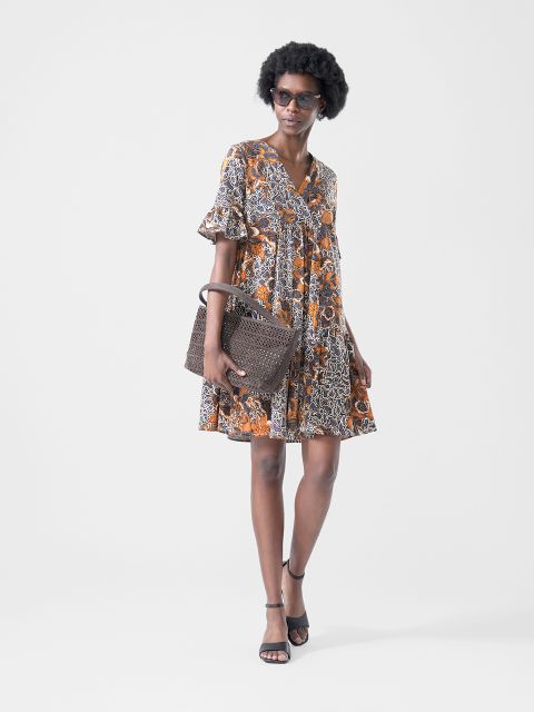 Mujer viste un vestido corto estampado en tonos naranjas y negros, gafas de sol, sandalias negras y lleva un bolso de mano marrón con textura.