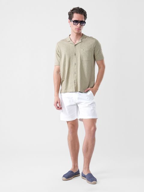 Un hombre con gafas de sol, una camisa de manga corta de color verde oliva claro, pantalones cortos blancos y alpargatas azul marino, de pie frente a un fondo blanco liso.