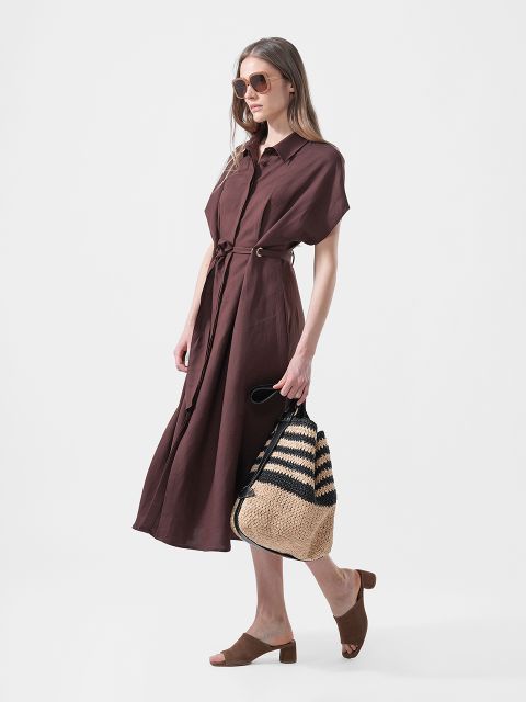 Una mujer lleva un vestido midi marrón con cinturón, sandalias planas marrones, gafas de sol y un bolso de paja trenzada con rayas negras.